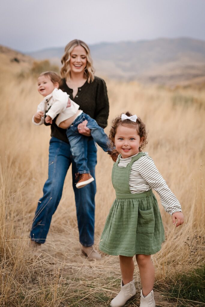 Mom holding baby while toddler daughter runs ahead smiling - candid family moment by Boise kids photographer Glean and Co in Boise Foothills setting