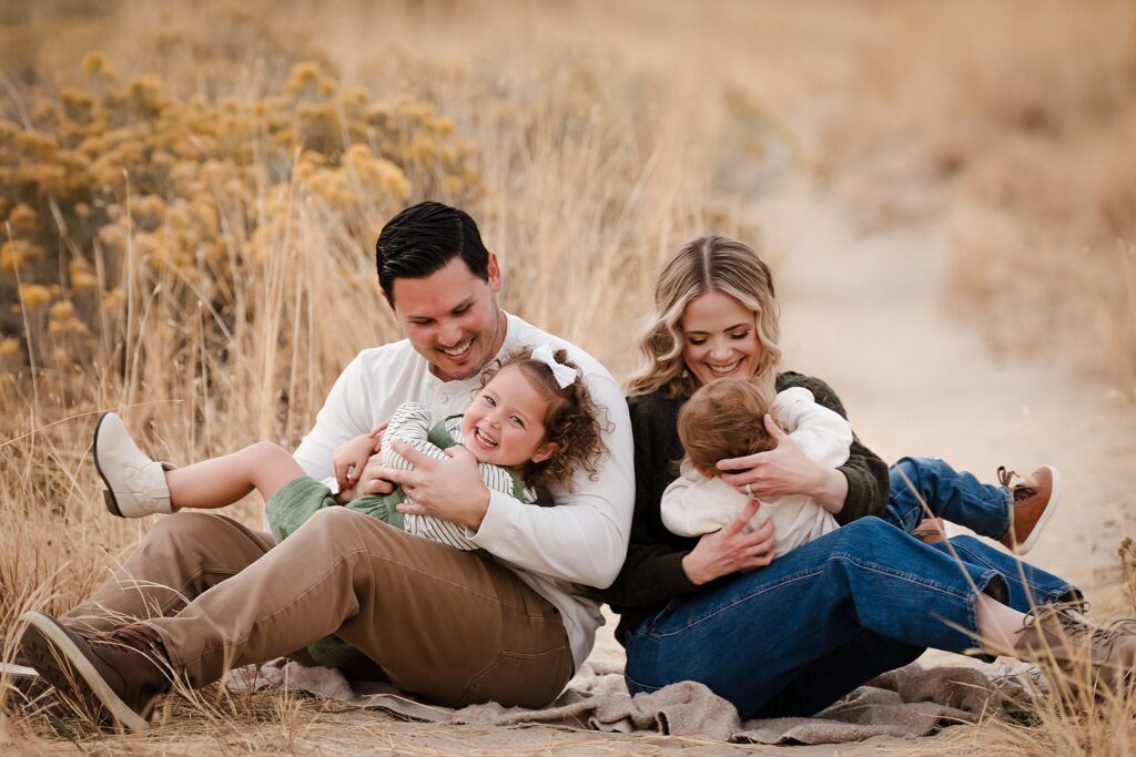Happy family of four laughing together sitting on blanket in golden grass field during Boise family portrait session