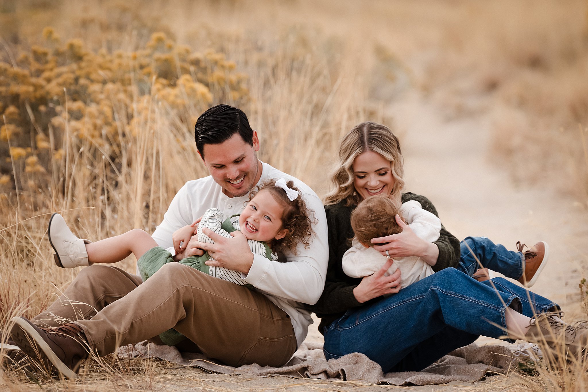 Happy family of four laughing together sitting on blanket in golden grass field during Boise family photo shoot