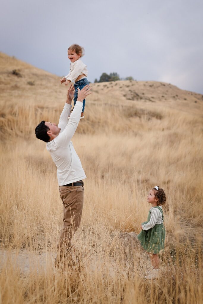Father lifting toddler boy high in the air while young daughter watches during Boise family photography session in golden field