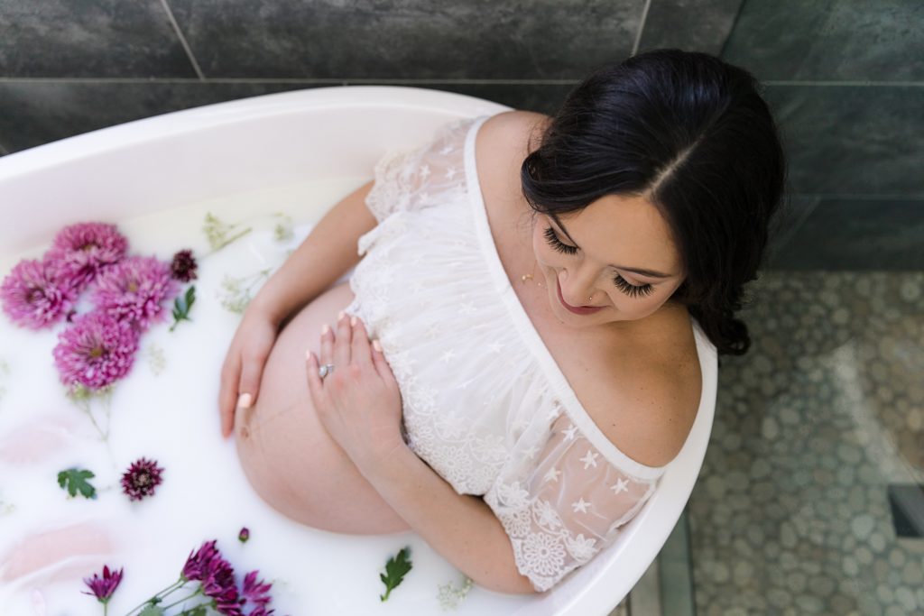 Pregnant woman in white maternity gown sitting in milk bath with purple flowers