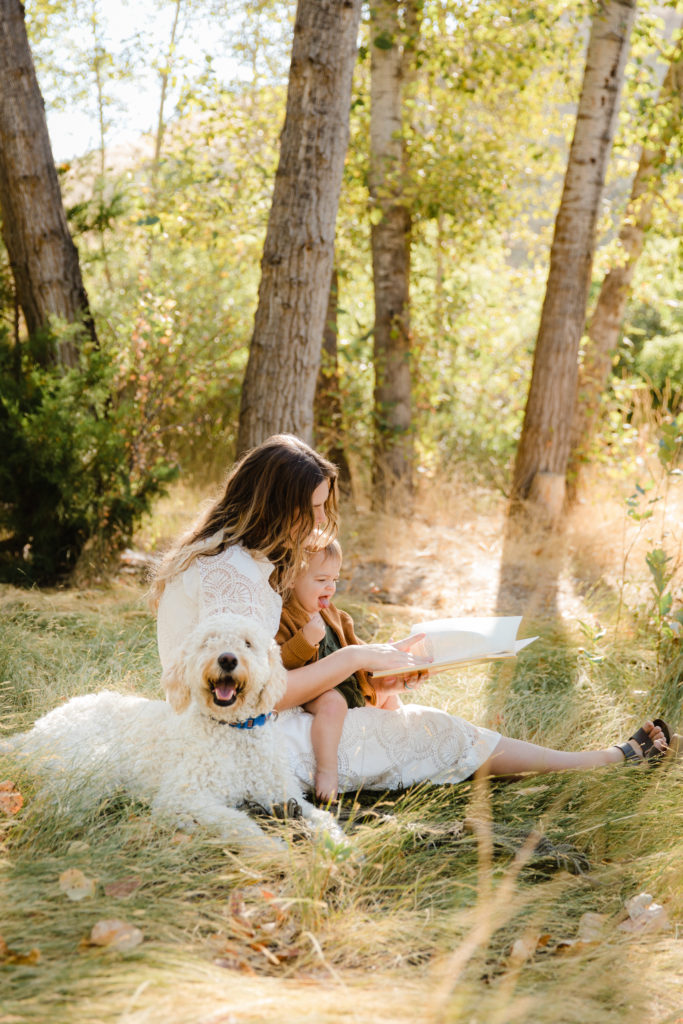 Mother reading to young child in dappled sunlight under trees with dog at her side