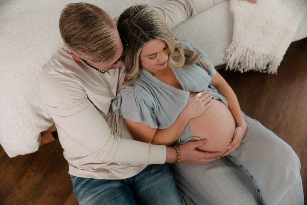 Husband and pregnant wife seated on floor leaning against bed and cradling baby bump