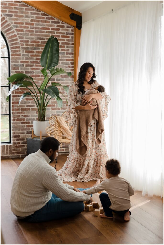 Mother holds newborn as she stands by a curtained window, with her husband and son seated on the floor nearby