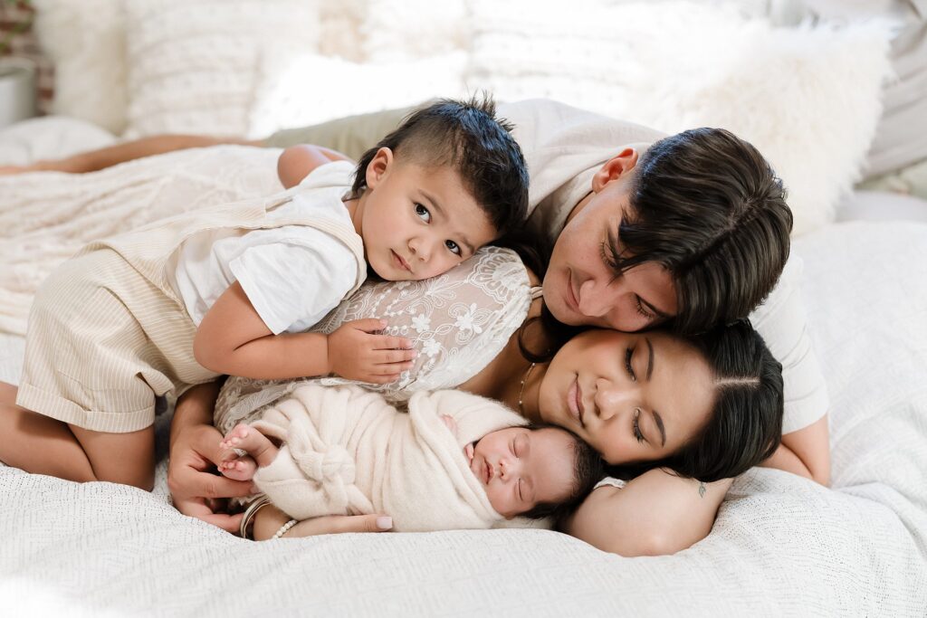 Family of four posing on bed, older son and dad hugging mom,  mom snuggling newborn