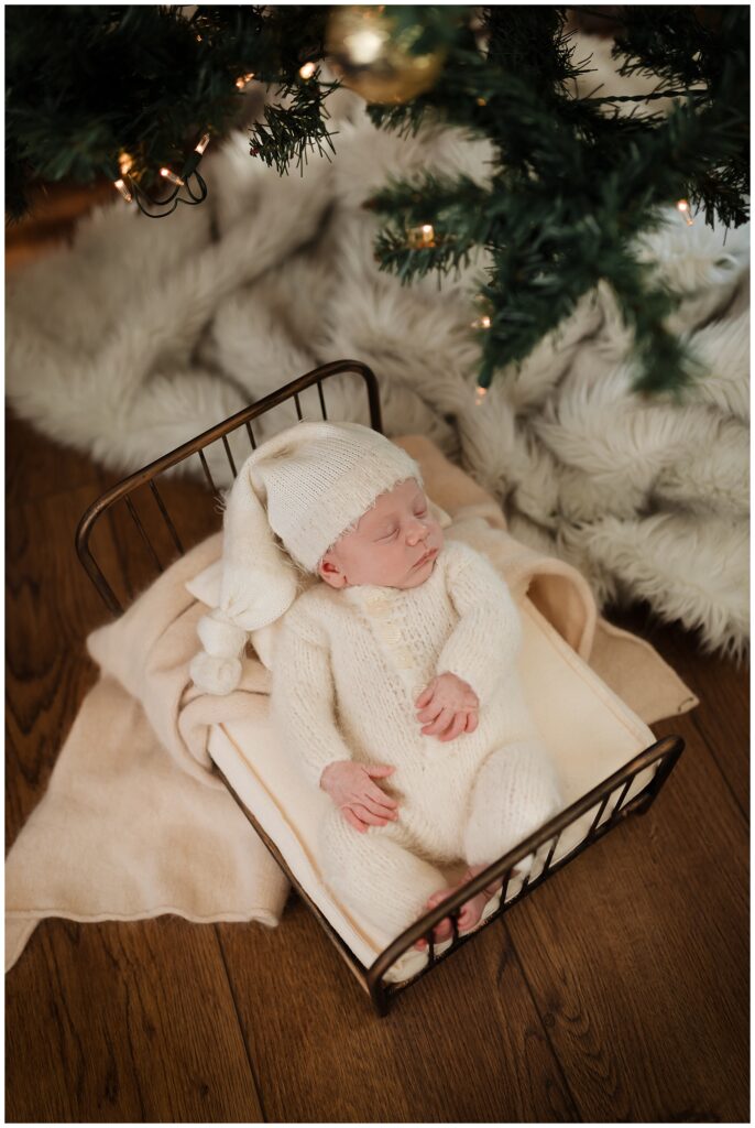 baby sleeping under a christmas tree wearing a fuzzy white romper and santa style sleepy hat