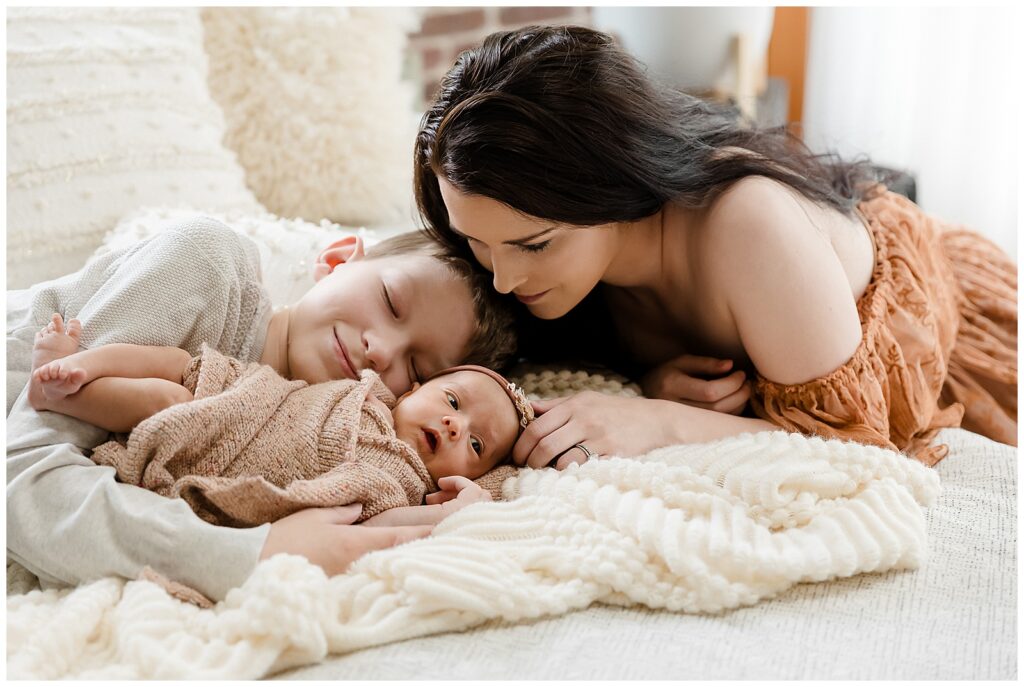 a mom in an off the shoulder lace gown gazes down at her newborn daughter while her older son cuddles his new sister