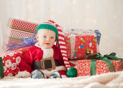 baby sitting among wrapped christmas presents wearing a santa costume and a long elf hat