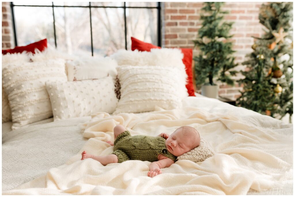 newborn baby boy in a green knot romber sleeping soundly on a cozy white bed surrounded by christmas trees