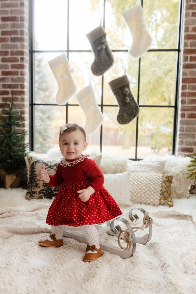 a toddler girl in a red dress celebrates the holiday season in front of a window of stockings in boise idaho