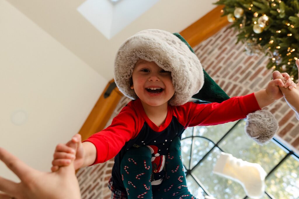 a young boy flies above his dads head in a game of airplane with a christmas tree in the background and a santa hat on his head, 