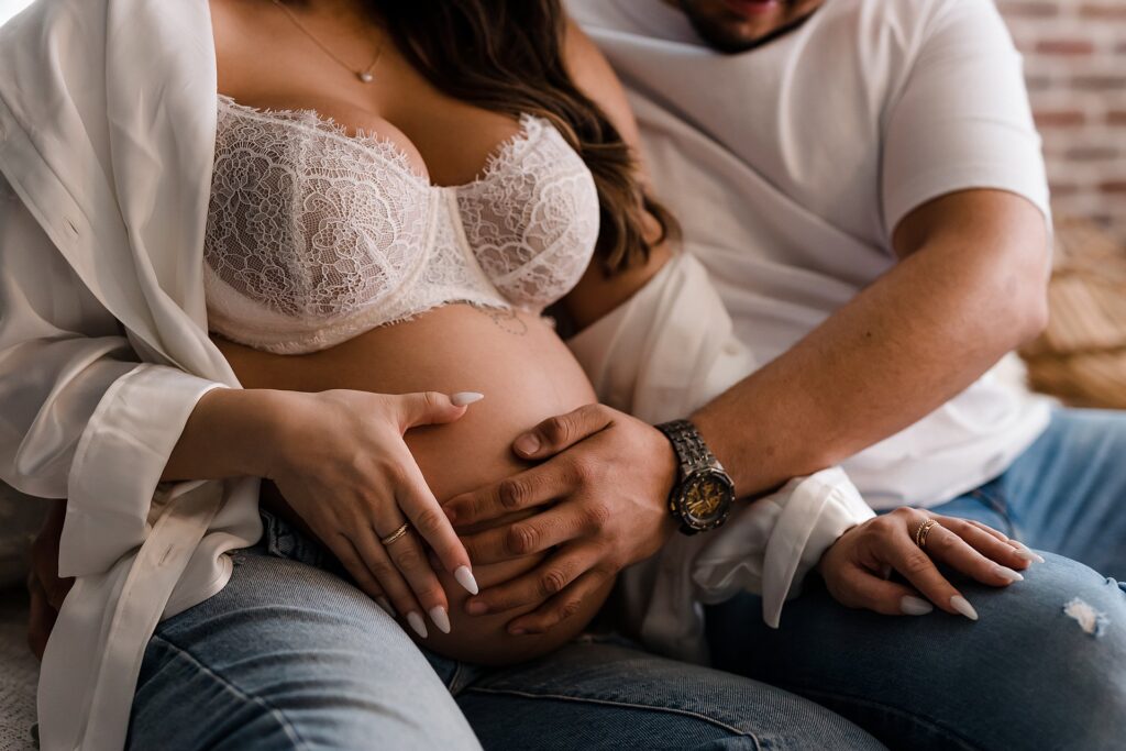 Intimate boudoir maternity portrait featuring white lace bralette and unbuttoned jeans, with father's hands tenderly placed on belly