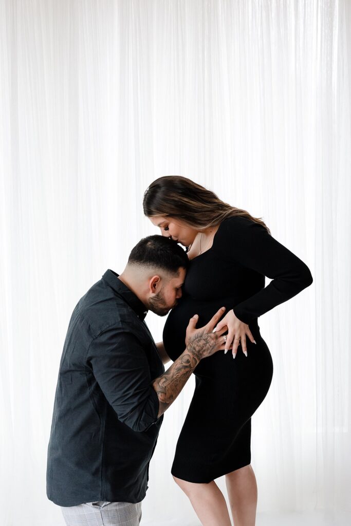 Elegant black dress maternity photo outfit captured against white curtains for timeless studio portraits