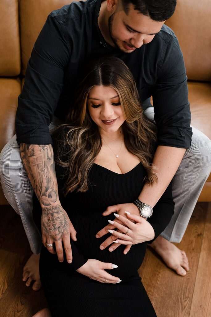 Contemporary maternity portrait of couple on leather couch, mother-to-be wearing sophisticated fitted black dress