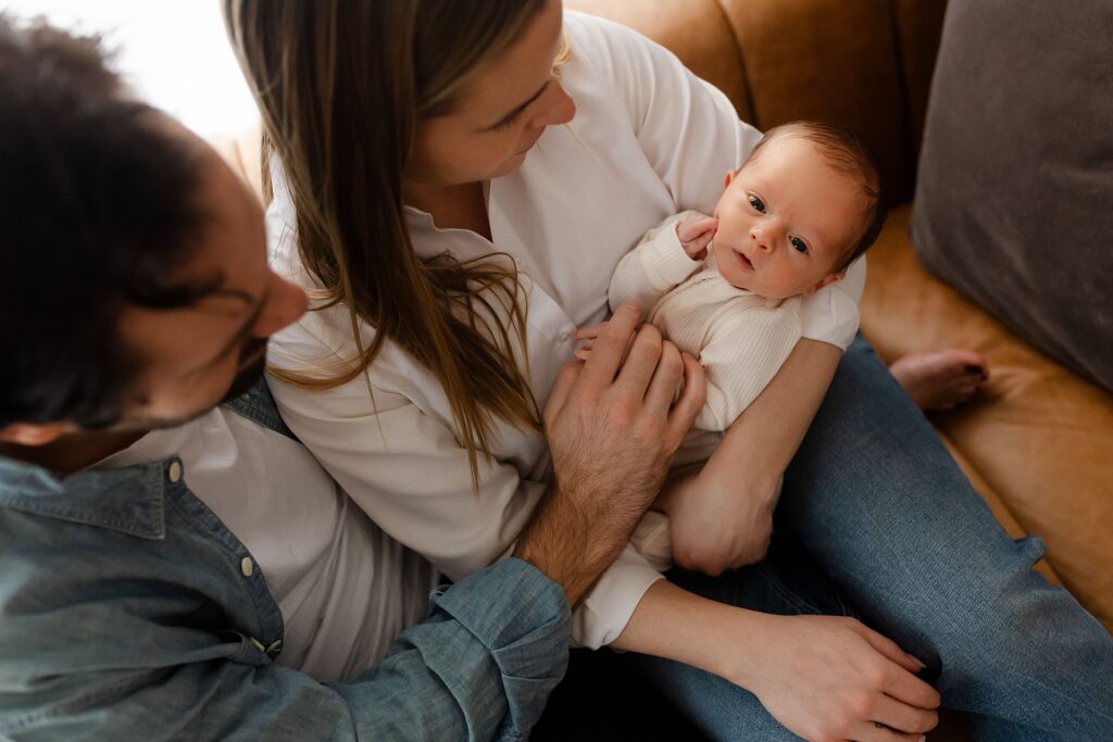 A heartwarming family portrait from above showing a baby boy looking directly at the camera while nestled in his mother's arms on their leather couch, his father gently holding his tiny hand with white curtains framing the scene