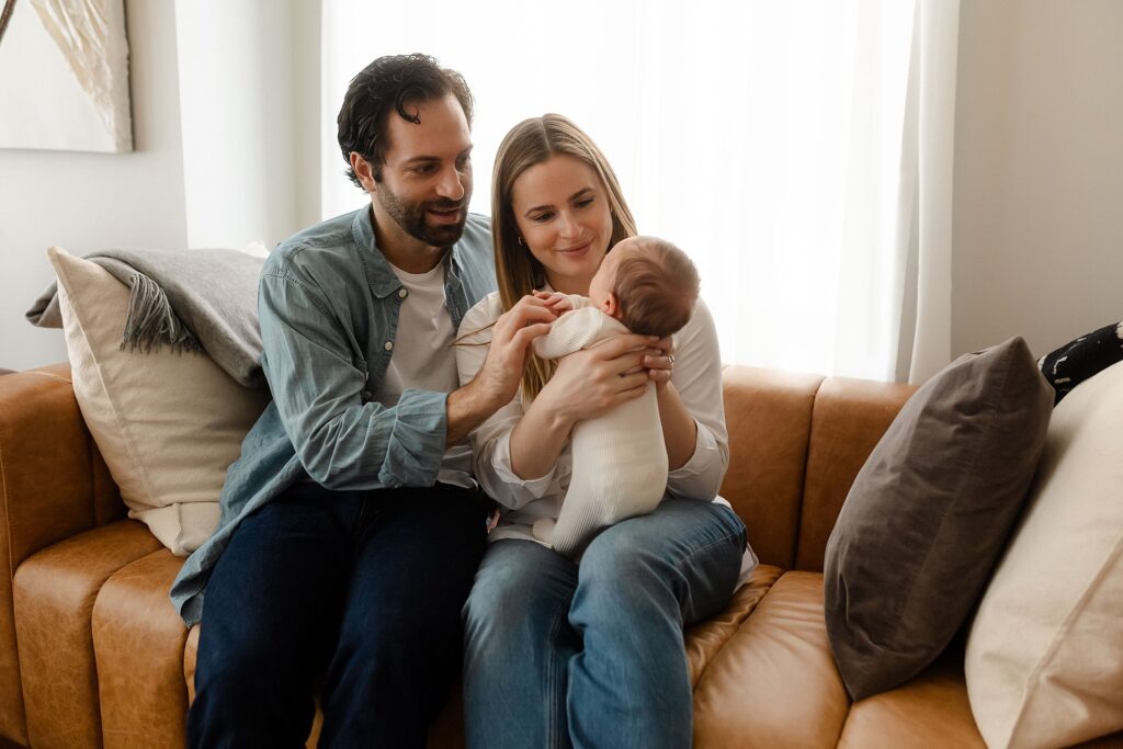 A heartwarming family portrait from above showing a baby boy looking directly at the camera while nestled in his mother's arms on their leather couch, his father gently holding his tiny hand with white curtains framing the scene