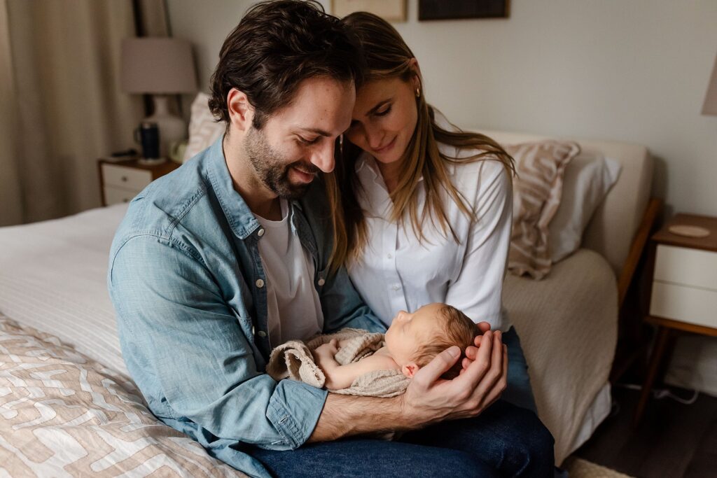 An intimate family moment showing a father cradling his newborn son while the mother gazes lovingly over his shoulder in their sunlit bedroom, capturing the tender support system essential for postpartum wellbeing