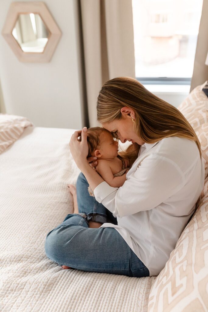 A tender moment between mother and newborn as she presses her forehead to her baby's, wearing a white shirt while sitting on their bed, illustrating the deep connection that can help navigate postpartum emotions