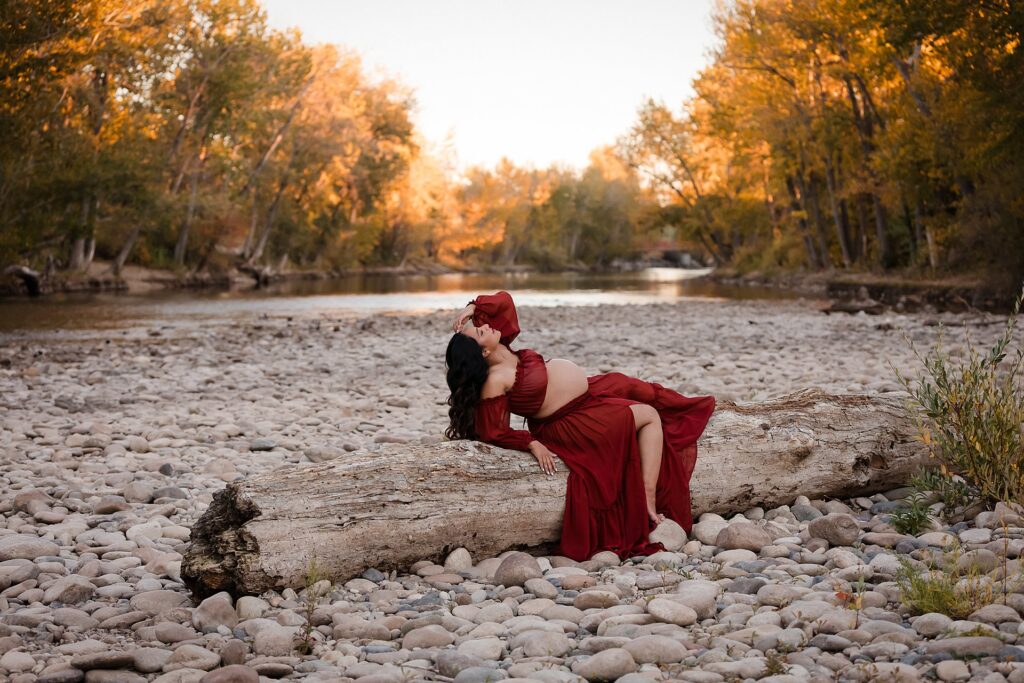 Expecting mother wearing a red two-piece maternity photo outfit poses elegantly on a log overlooking the Boise River during sunset