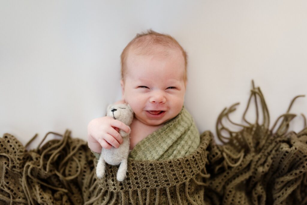 a baby boy smiling, holding a teddy bear during his newborn photography session.