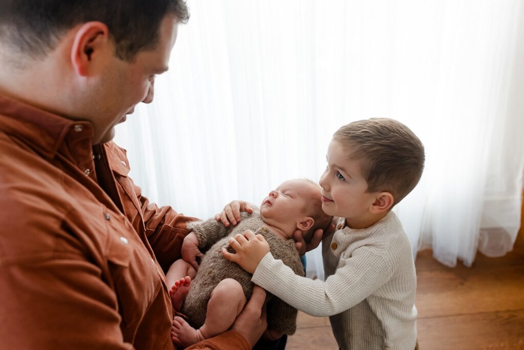 a dad holding a newborn baby out so that his older son can snuggle on his new sibling for a photo together