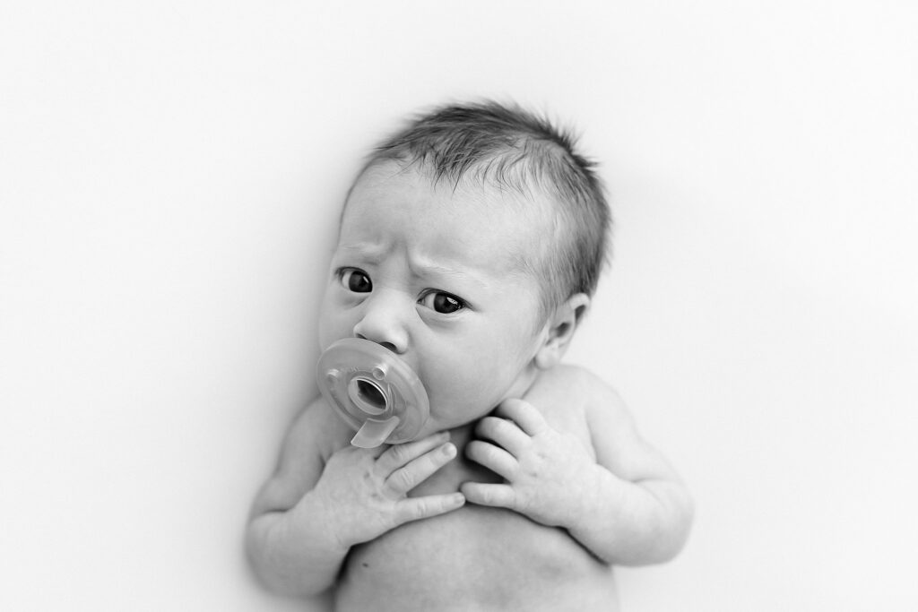 a newborn baby with a pacifier looks at the camera suspiciously, perhaps wondering what photo retouching is going to happen to her photo.