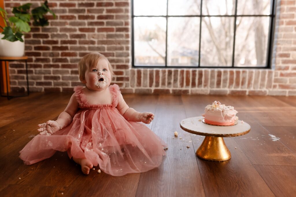 One-year-old girl in a fluffy pink tutu dress excitedly smashing her pink birthday cake on a gold stand, sitting on a wood floor against a brick backdrop as frosting covers her hands and cheeks.