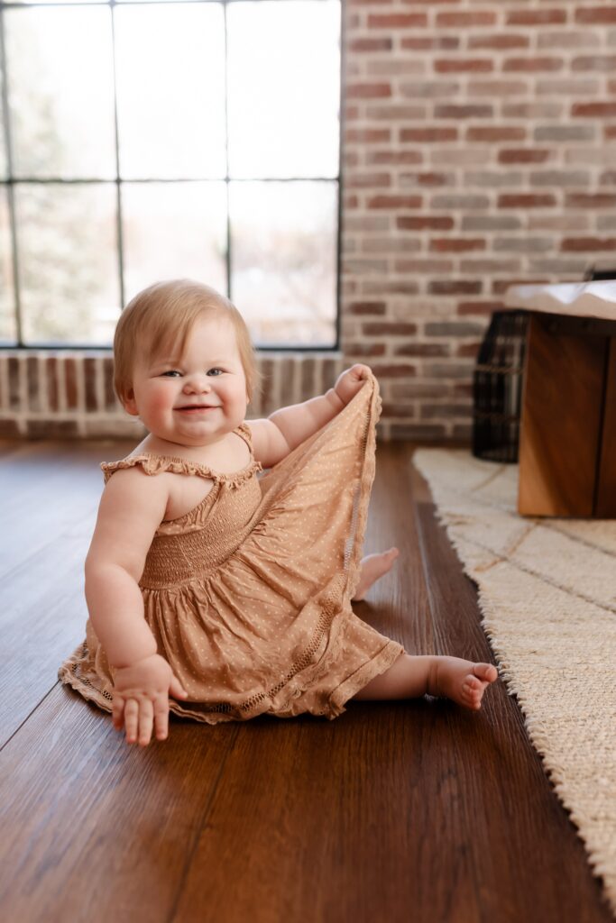 One-year-old birthday girl posing sweetly in her pre-cake pink tutu on the wooden floor with brick wall behind her, showcasing her personality before the messy pink cake smash adventure begins