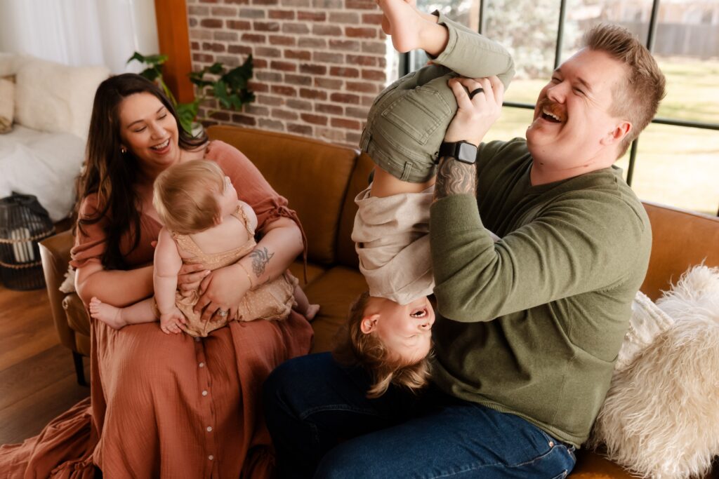 "Playful family moment before the cake smash - dad holding his son upside down with laughter while mom snuggles their daughter in her pink tutu against the brick wall and wooden floor, capturing the family's joyful energy before celebrating