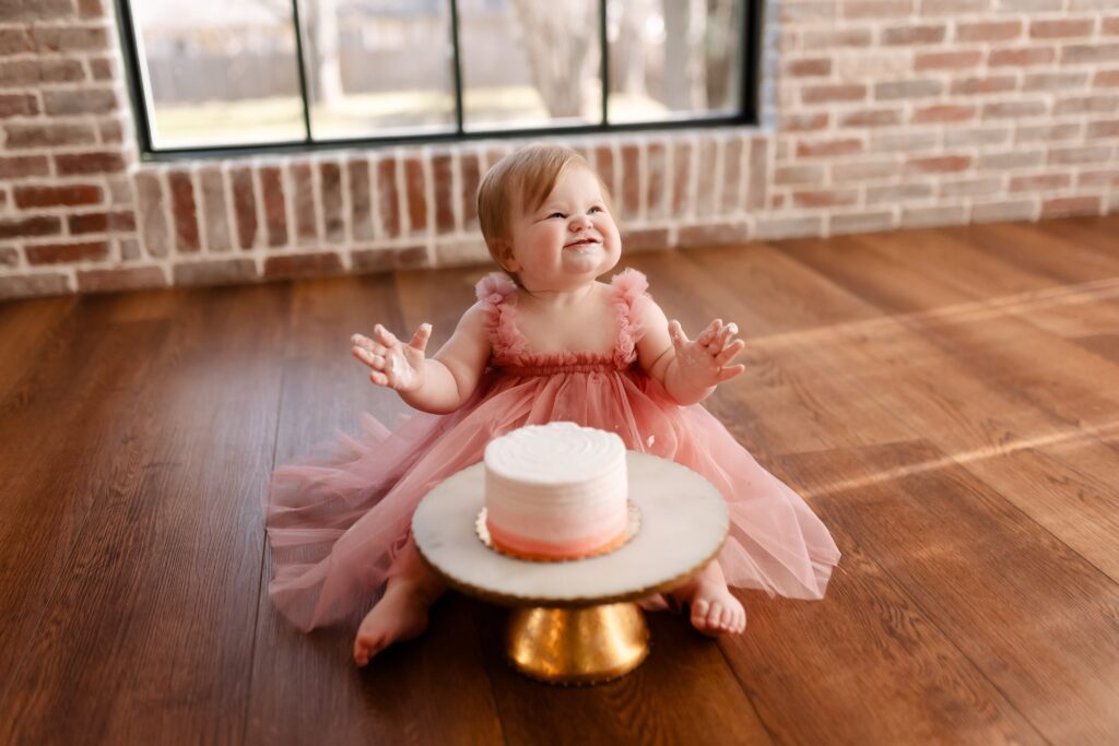 a one-year-old in a pink tutudress fully embraces her cake smash session, pink frosting splattered across the wood floor and gold cake stand, her laughter captured against the rustic brick wall background.