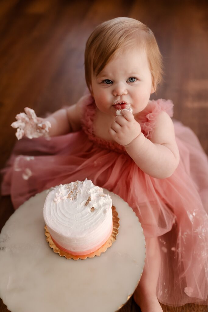 Curious one-year-old in a pink tutu dress brings her frosting-covered finger to her lips for the first taste of her birthday cake, her expression full of wonder as she sits on the wooden floor with the partially smashed pink cake on its gold stand