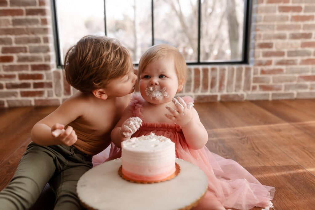 Brother and sister cake smash moment on wooden floor - big brother kissing his little sister in pink tutu as she digs into the pink birthday cake, creating sweet sibling memories against the brick wall backdrop