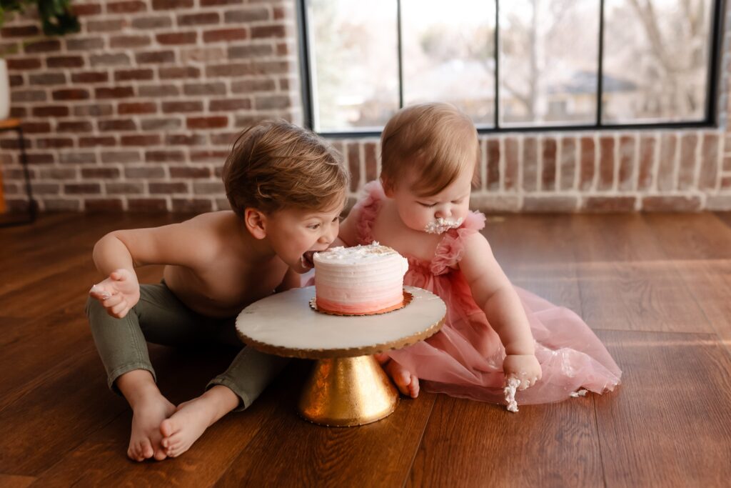 Big brother leans in for a hands-free bite of the pink cake while his little sister focuses on painting patterns with her frosting-covered finger on the wooden floor, each exploring the cake smash experience in their own unique way