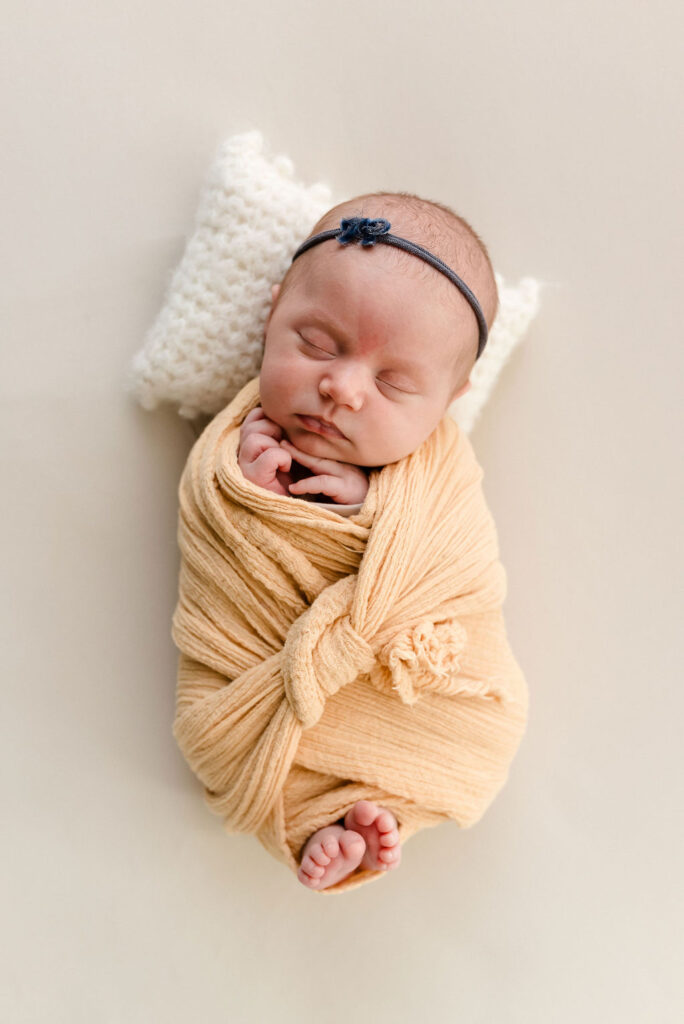 newborn baby girl in a yellow wrap with a tiny blue headband and white pillow behind her head.