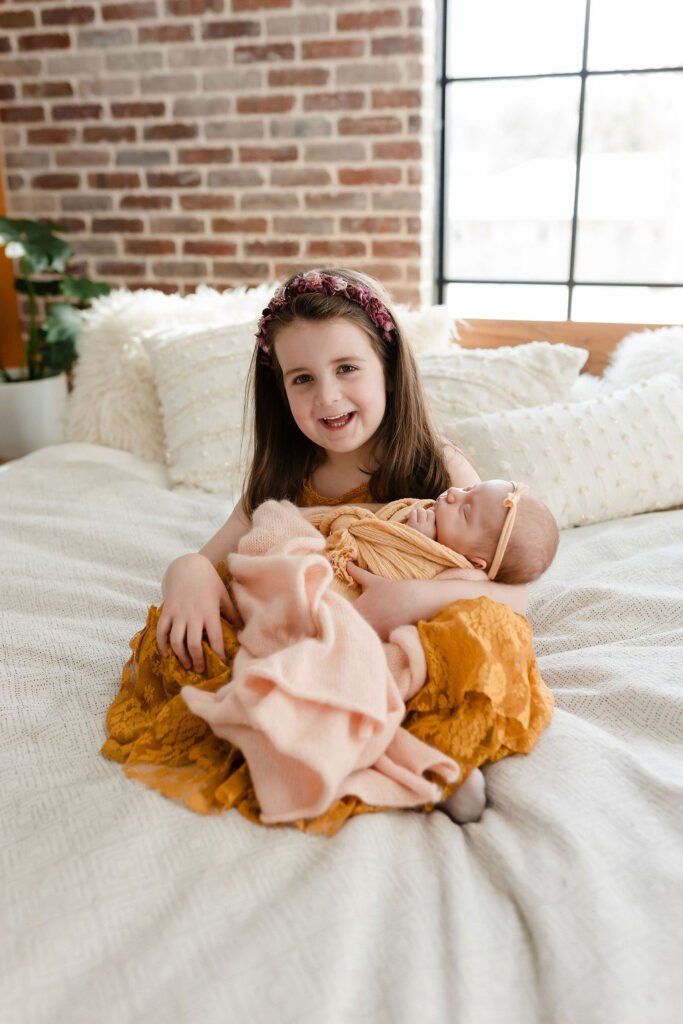 big sister in a yellow dress holds her new baby sister during a lifestyle mini newborn session