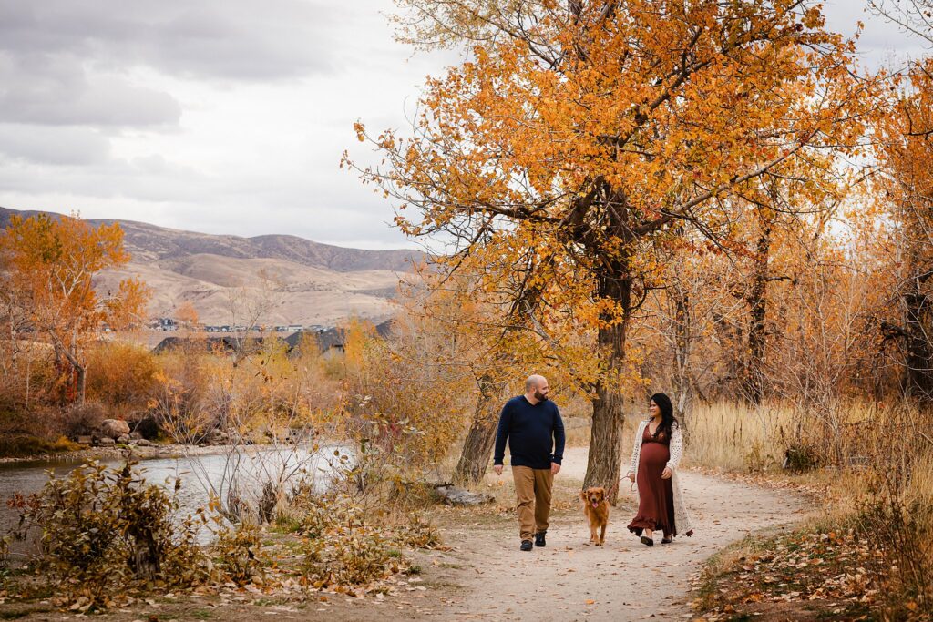 Couple with dog walking on autumn greenbelt during twin maternity photo shoot in Boise Idaho"