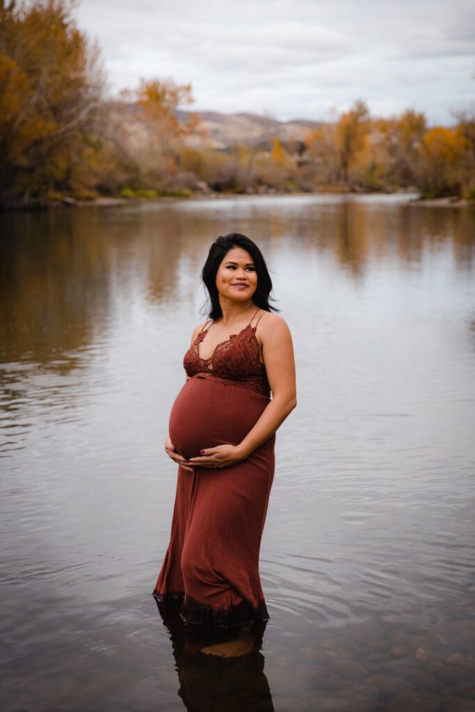 Pregnant woman in burgundy dress standing in the Boise River during twin maternity photography session