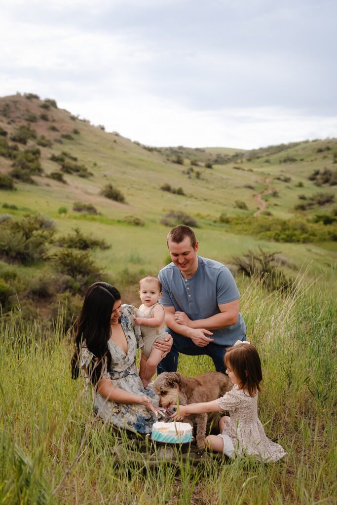Family photography with dogs including small dog during toddler birthday celebration with parents, baby, and birthday cake in grassy Boise area field location