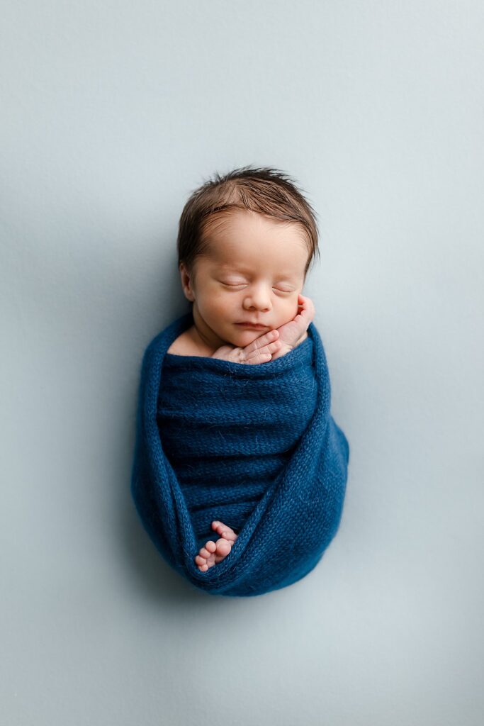 a newborn baby boy wrapped in a dark blue wrap with a light blue background, the opposite of his twin brother