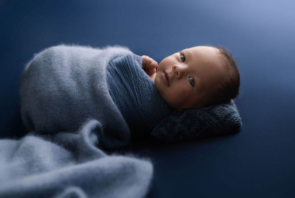 Side-lying newborn baby in blue knit wrap on textured backdrop