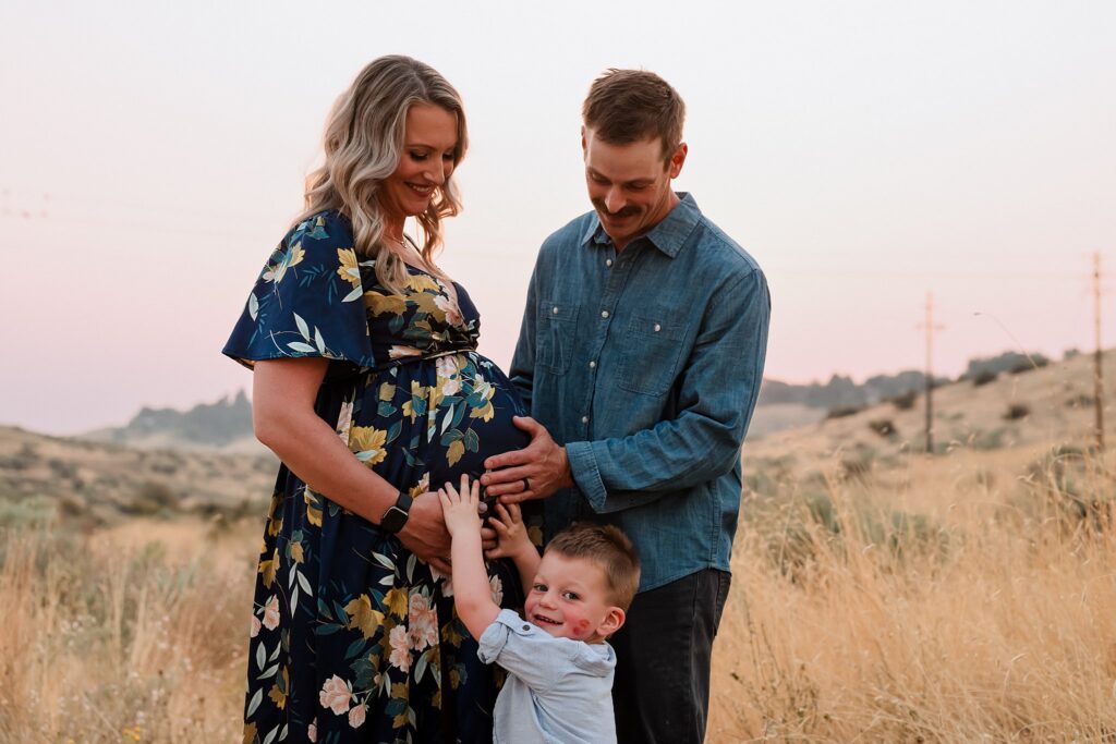 Pregnant mother, father, and toddler son together in sage brush field during Boise location scouting for family photos maternity session at golden hour