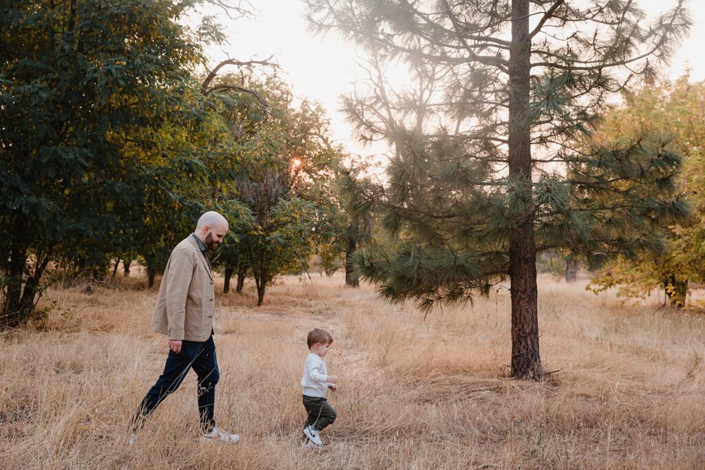 Dad and young child playing together in dappled shade of trees during professional Boise location scouting for family photos session with natural lighting