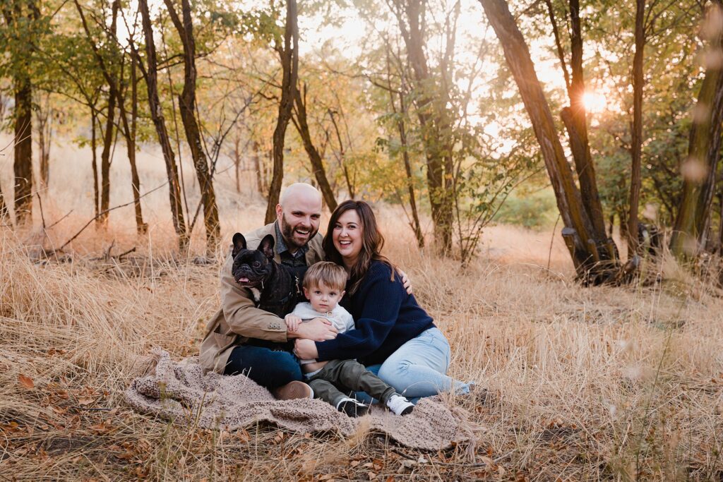 Family of three sitting on blanket under filtered sunlight through trees during Boise location scouting for family photos session in wooded area behind Foothills Learning Center