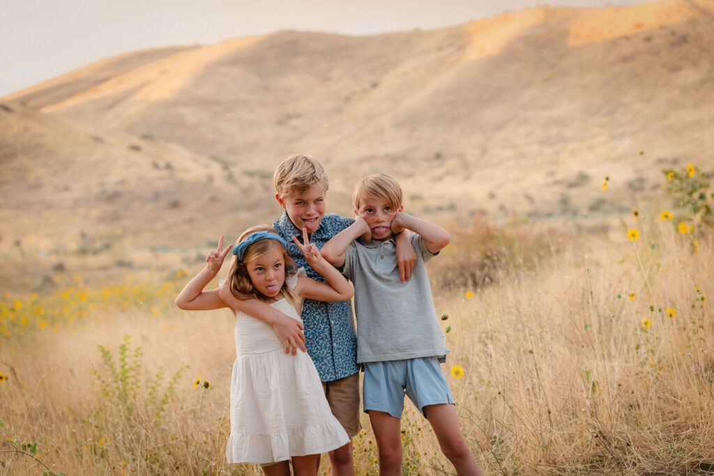 Three children playing in wild sunflowers with foothill views during sunflower family photos Boise Idaho session by top Boise family photographer