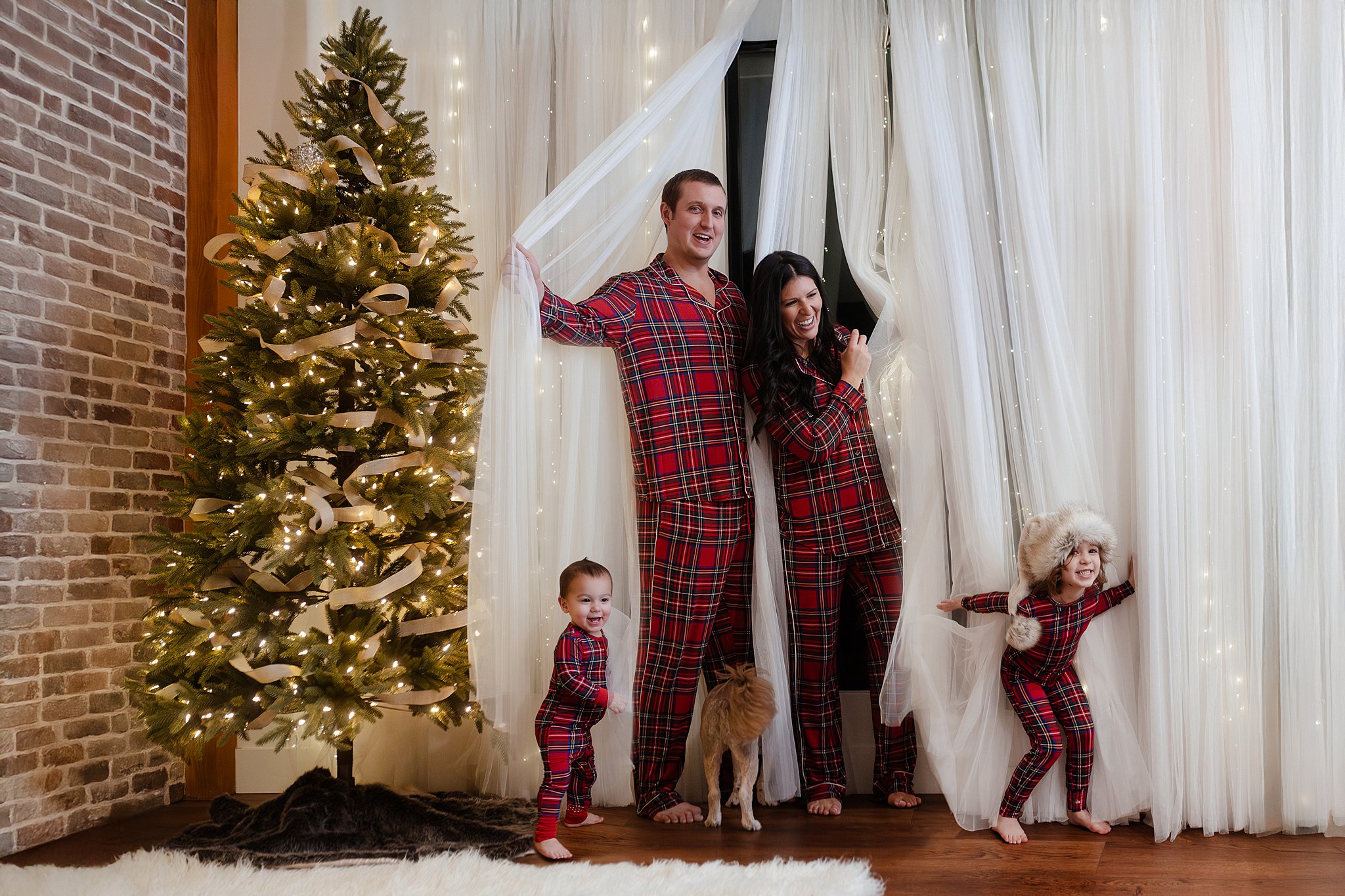 a family of 4 peeking out of holiday curtains in the Glean and Co photography studio for their annual matching pajamas holiday photos